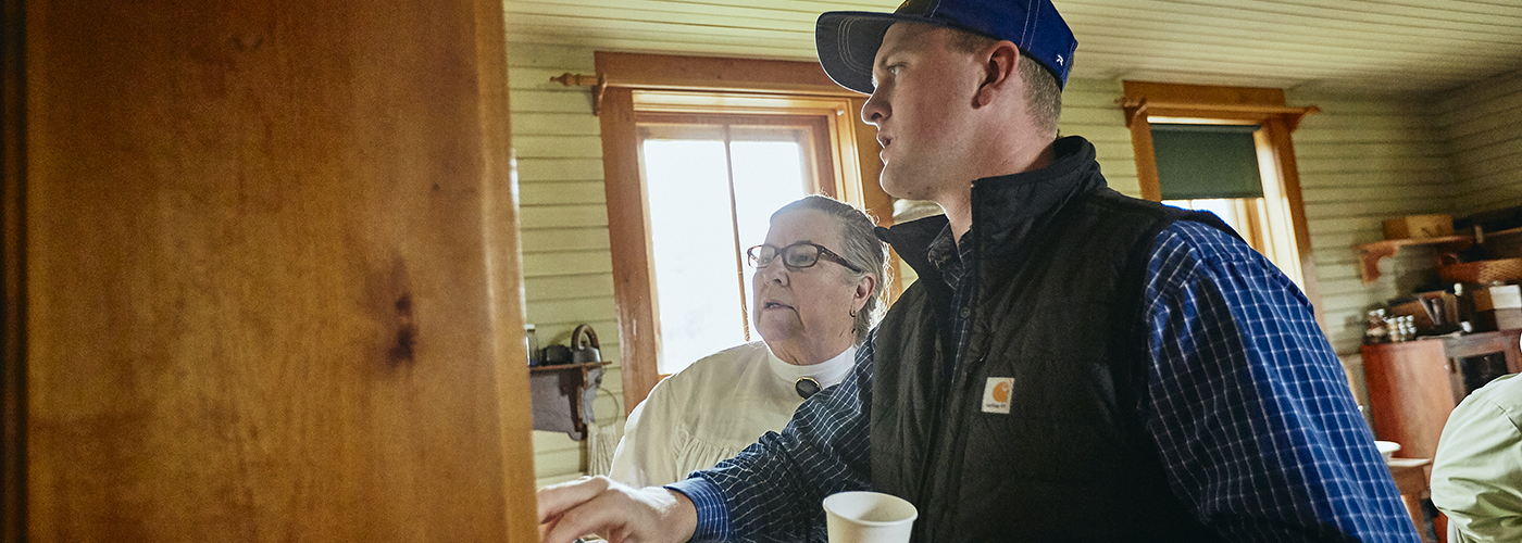 A young Caucasian man with a ballcap on discusses an artifact with a shorter, older woman in frontier garb of a blouse and skirt. Placeholder image