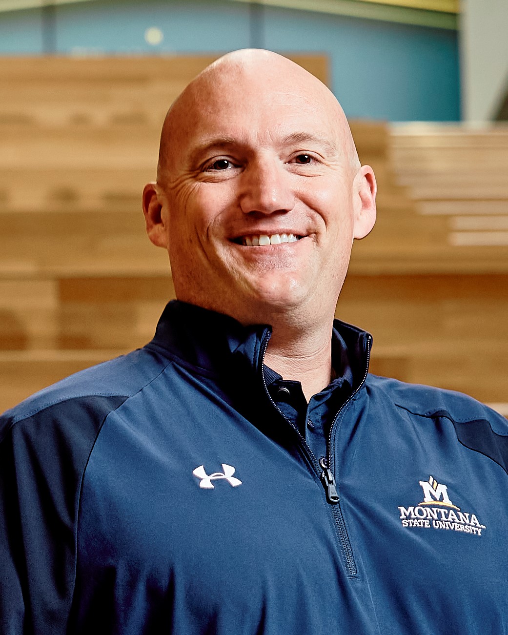 Brock LaMeres photo of man with a bald head and brown eyes wearing a blue Montana State University logo shirt, standing in front of wooden steps