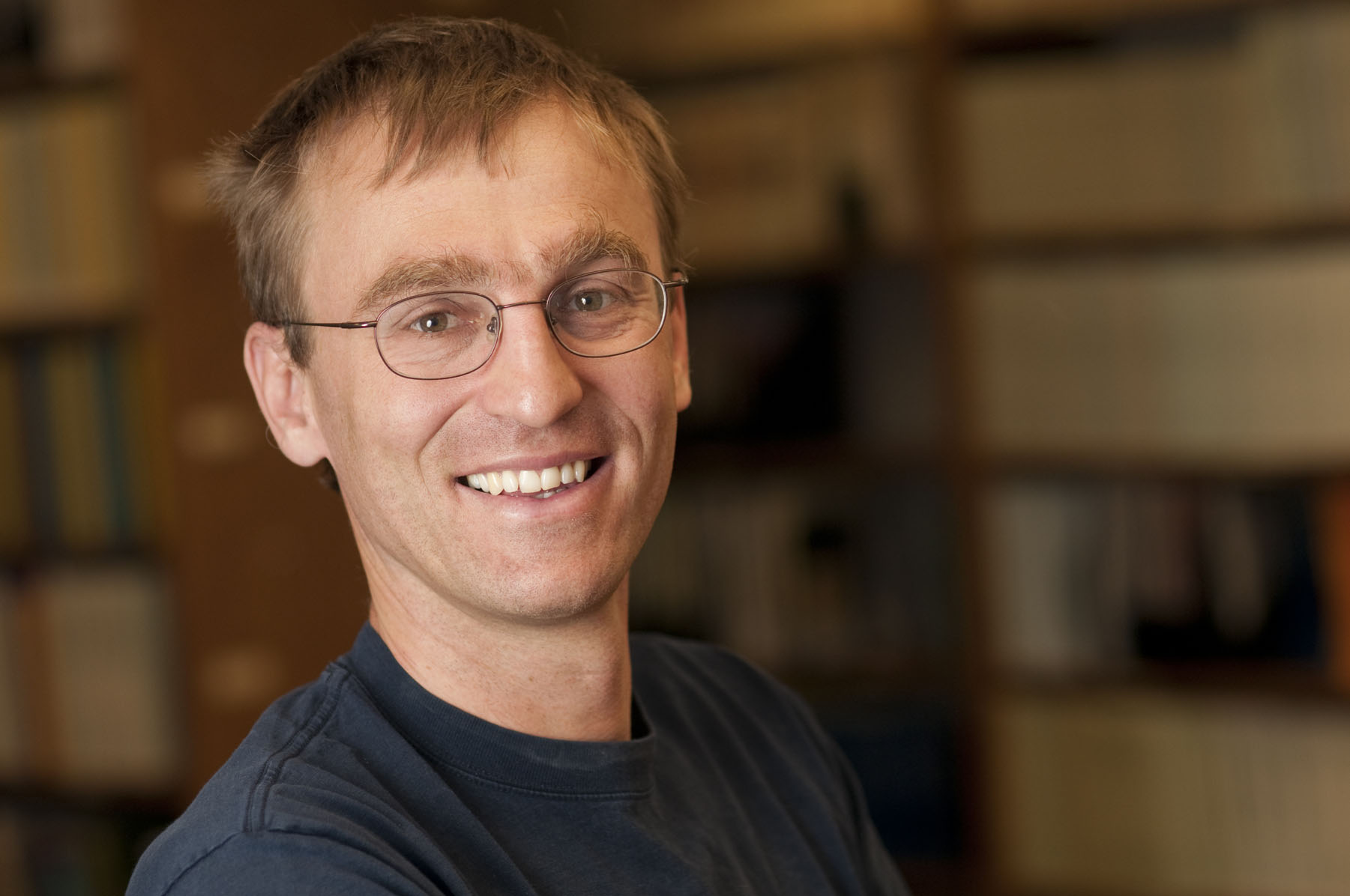Tomas Gedeon Man with short brown hair and glasses wearing a blue shirt standing in front of a blurred out background of books on a shelf