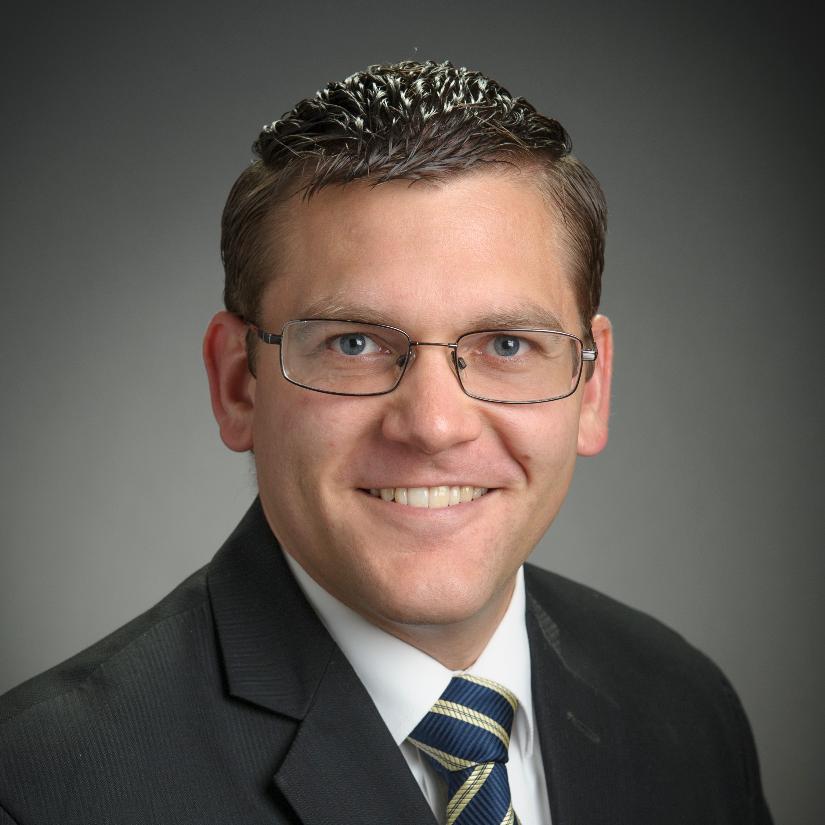 Bradley Whitaker photo Caucasian man with short dark hair wearing a suit, tie and glasses with a gray background.