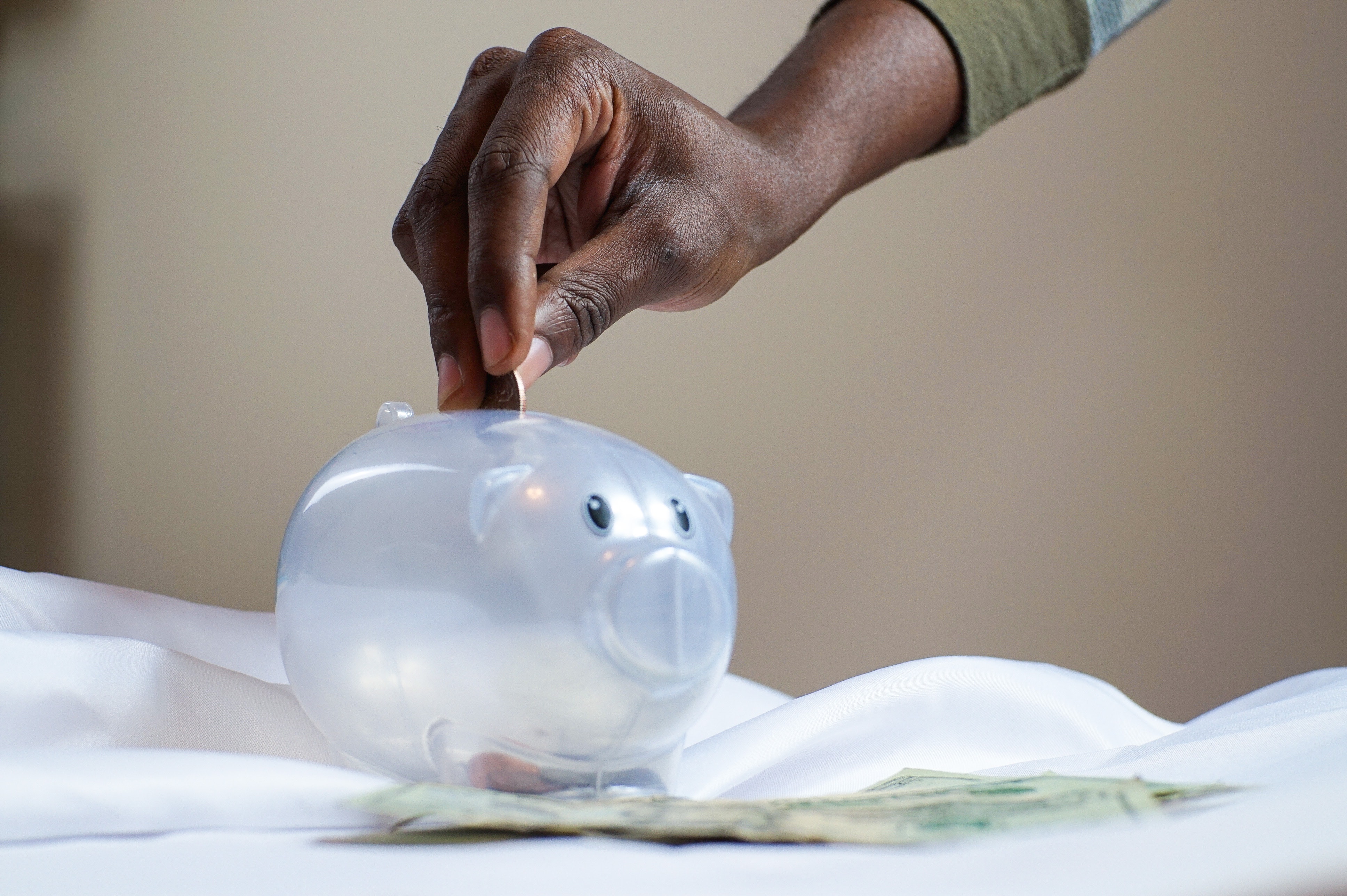 Saving Money hand adding coin to clear piggy bank sitting on a white table cloth.