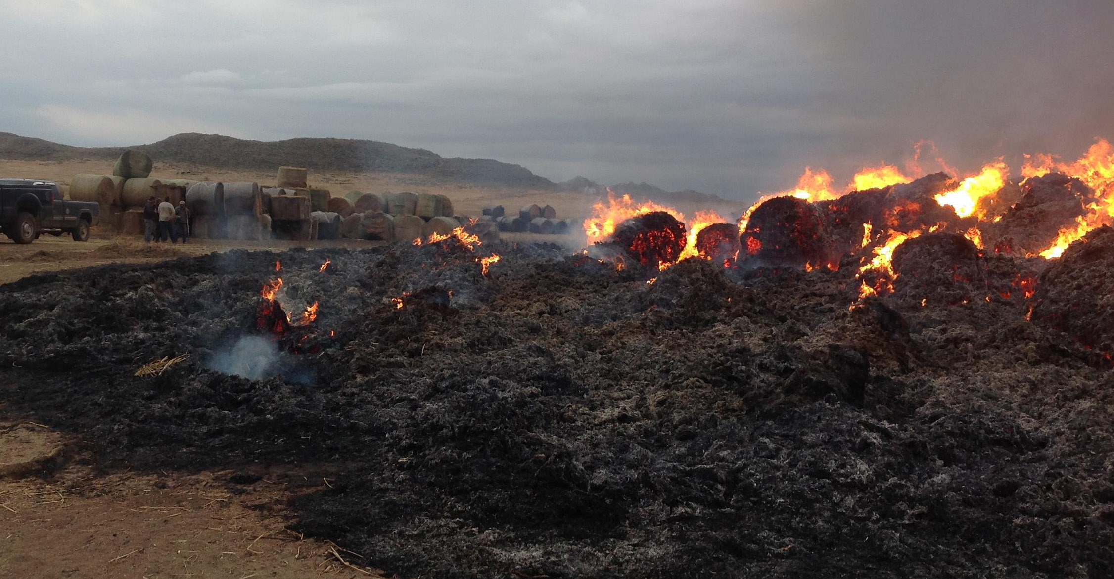 Several round bales of hay are black and on fire.