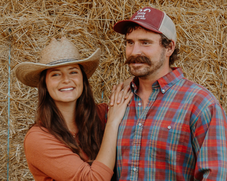 A man and woman, Caroline and Justin Nelson, stand together in front stacked hay bales. 