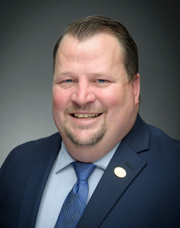 Cody Stone Headshot Cody Stone in a navy suit jacket with a light blue button up and blue tie on. There is a circular, gold pin attached to his left suit lapel.