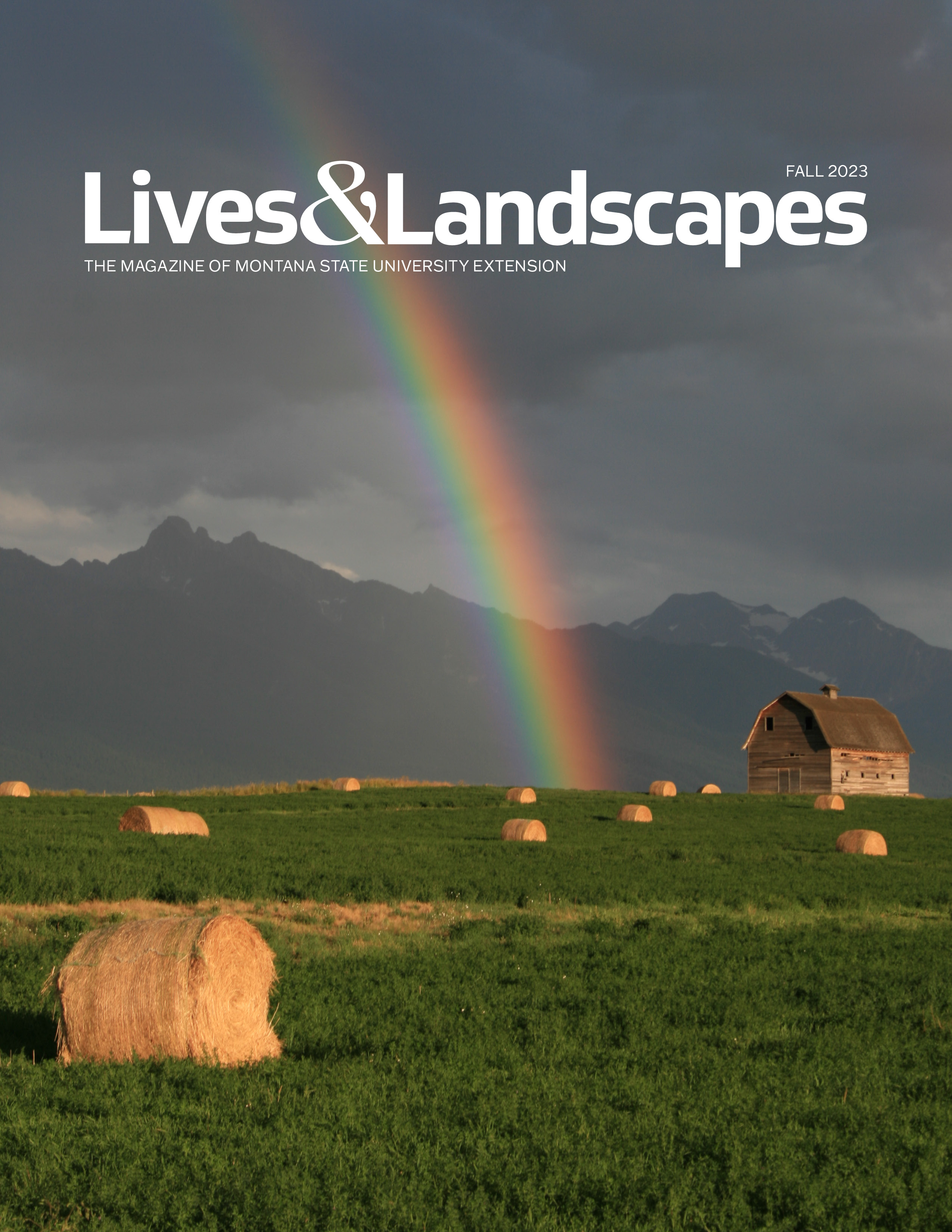 A bright rainbow emerges from a dark grey sky and lands in a green field dotted with haybales that are lit up by the sun. A barn sits to one side and there are mountains in the distance.