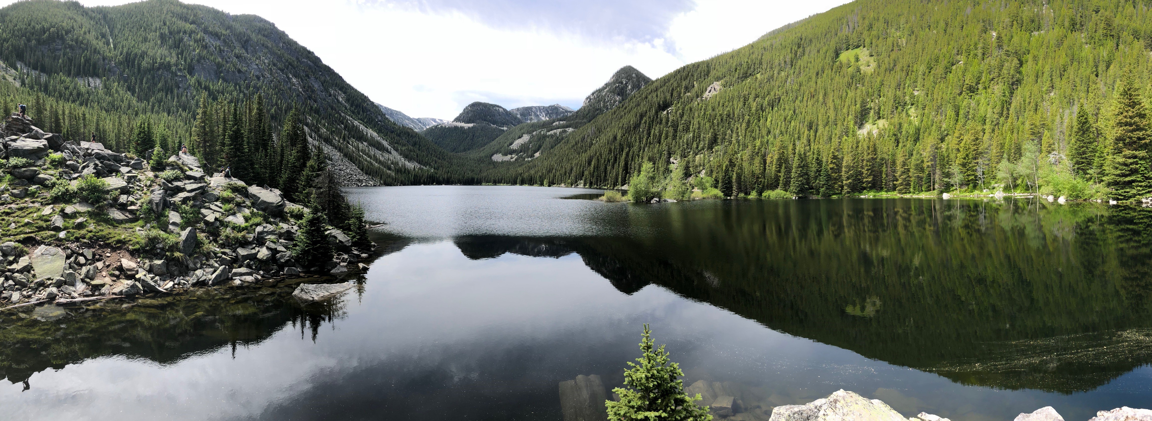 Lava Lake, MT by retired test center coordinator, Rick Jackson Panoramic view of a serene mountain lake surrounded by lush green forests. The lake reflects the surrounding landscape, creating a mirror image on its surface. Large rocks are visible on the left side of the lake’s shore, and the forested mountains enclose the lake in a tranquil embrace.