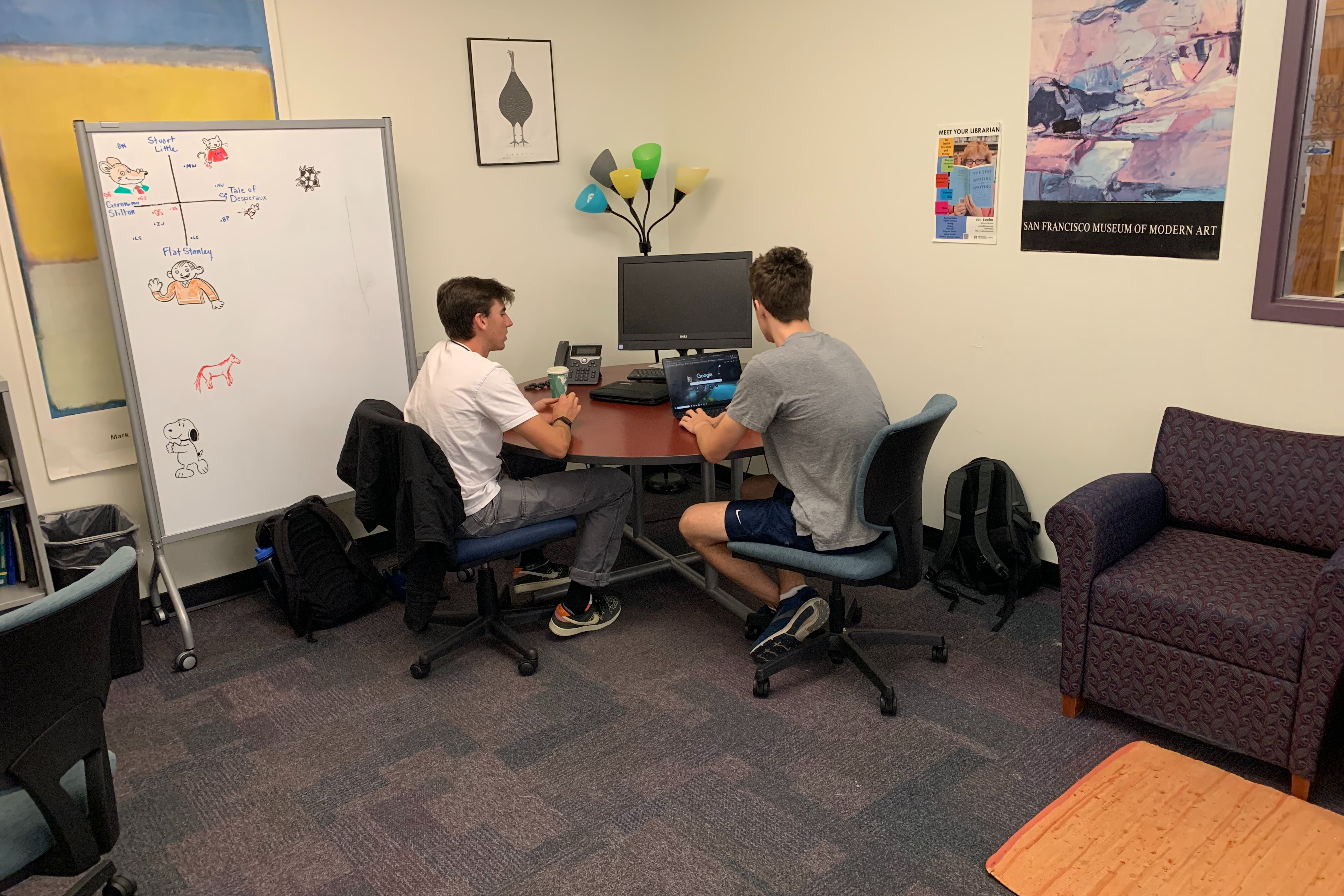 A writer and a tutor working together at a small table in the corner of the Library Writing Center. There is a whiteboard covered in notes in the background.
