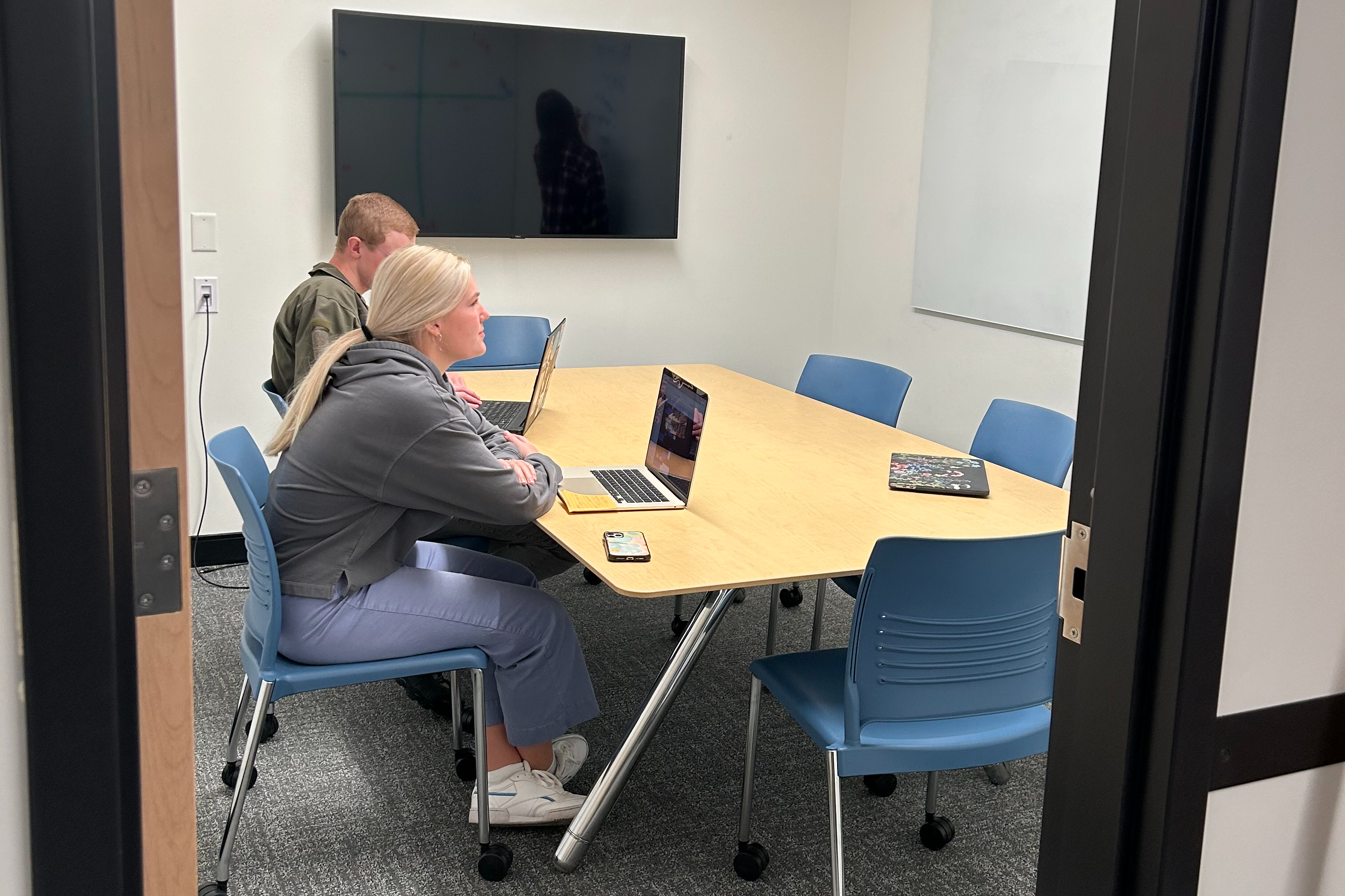 One of the Conference Rooms in the Romney Writing Center, with table and six chairs, as well as a large TV screen. Two writers sit at the table working. 
