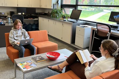Two writers sit on comfortable orange couches in the Wilson Writing Center, reading. Natural light streams in through the windows nearby.